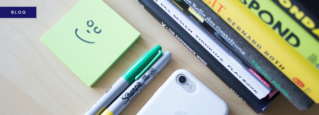Blog banner showing two Sharpie pens, an iPhone and a pile of books on a desk, next to a post-it pad with a smiley face drawn on the top post-it